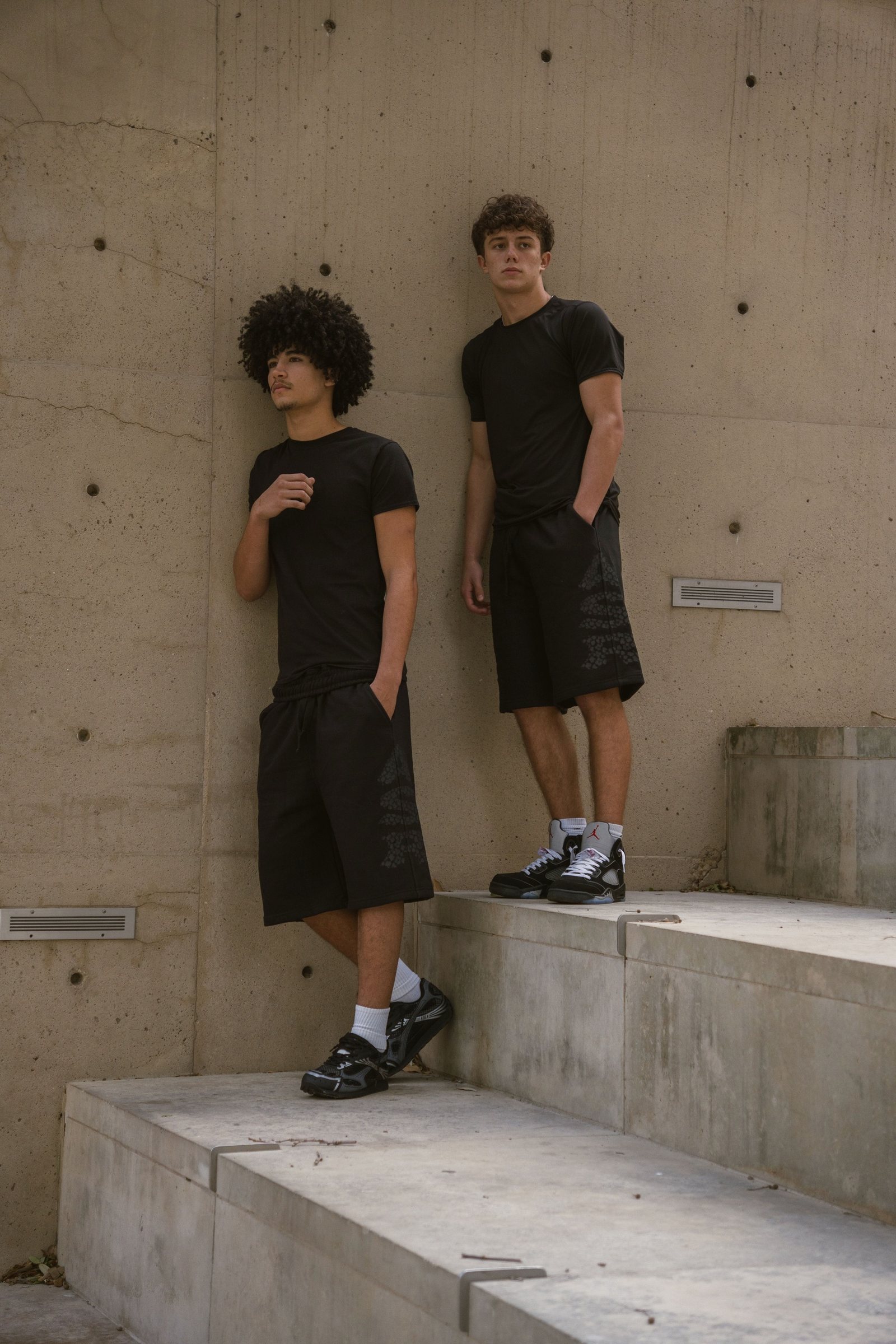 Two models in Asior tees and sweat shorts on concrete steps