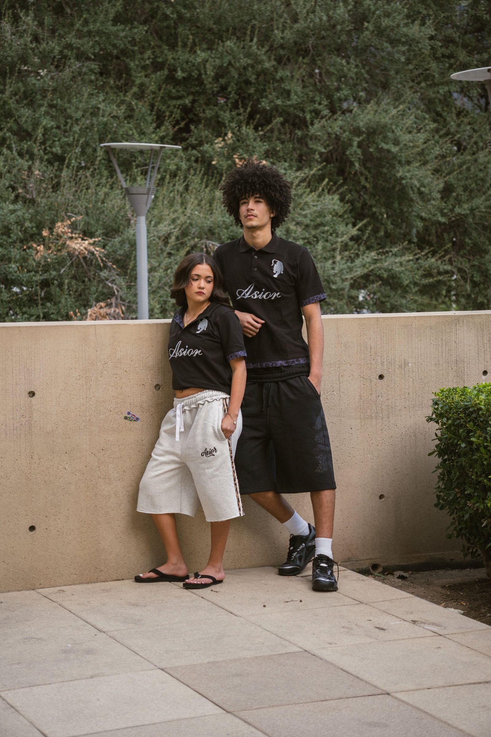 Two models in Asior polos and sweat shorts against a concrete wall